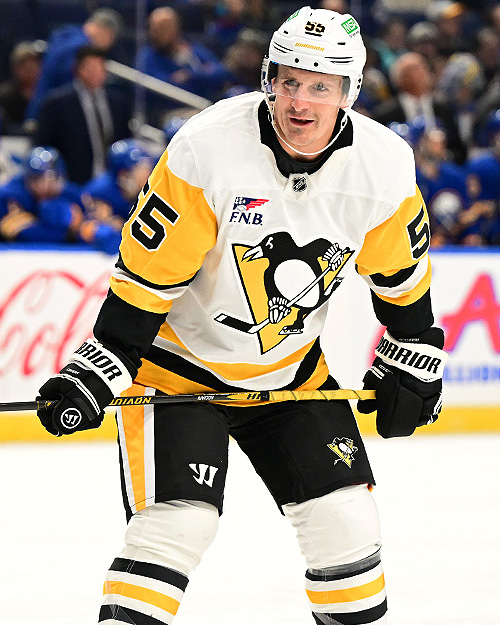 A hockey player wearing a white and yellow jersey with the logo of the Pittsburgh Penguins, holding a hockey stick and smiling on the ice during a game.