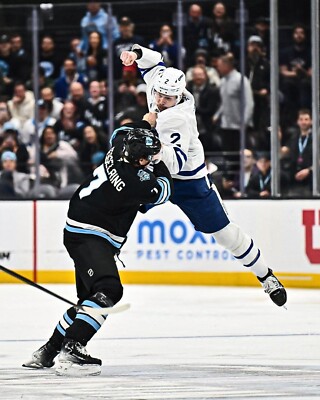 A dynamic moment in a hockey game, featuring a player in a white jersey leaping in the air while engaging with an opposing player in a black jersey.